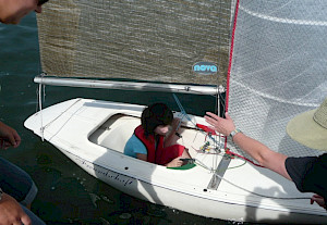 Ein Kind sitzt in einem kleinen Segelboot und ein Erwachsener hält es vom Steg aus an einer Leine fest.
