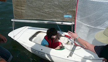 Ein Kind sitzt in einem kleinen Segelboot und ein Erwachsener hält es vom Steg aus an einer Leine fest.